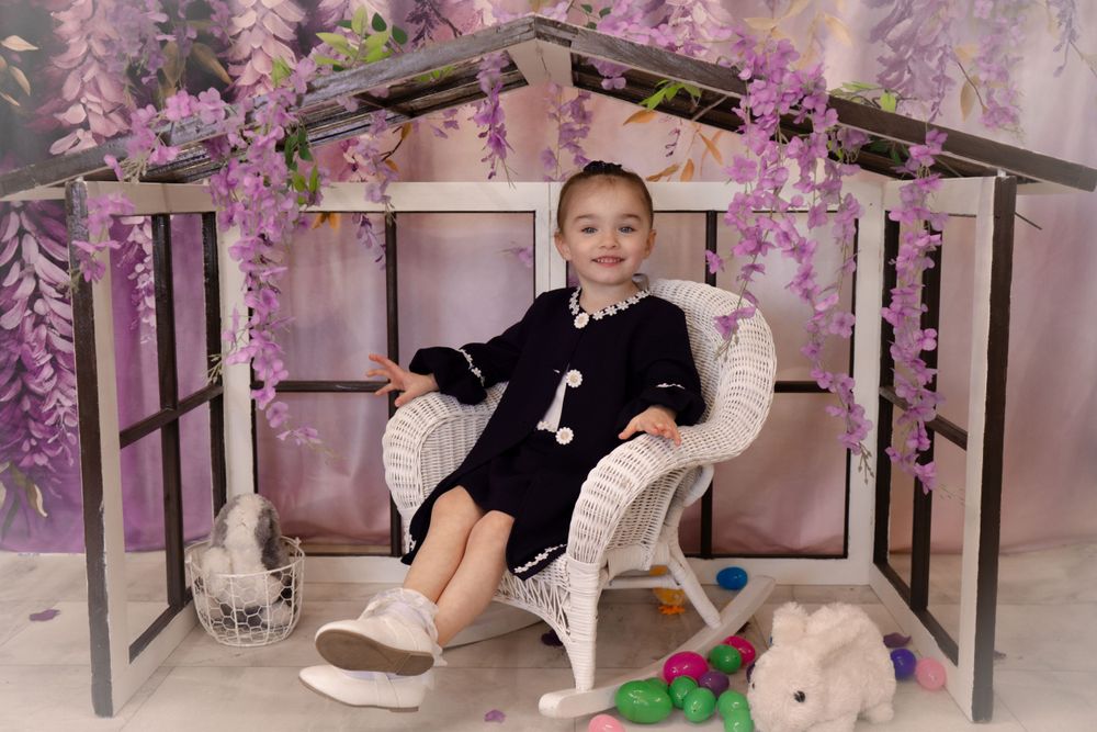 A stylishly dressed toddler sits in a white wicker chair surrounded by purple flowers and plush toys.