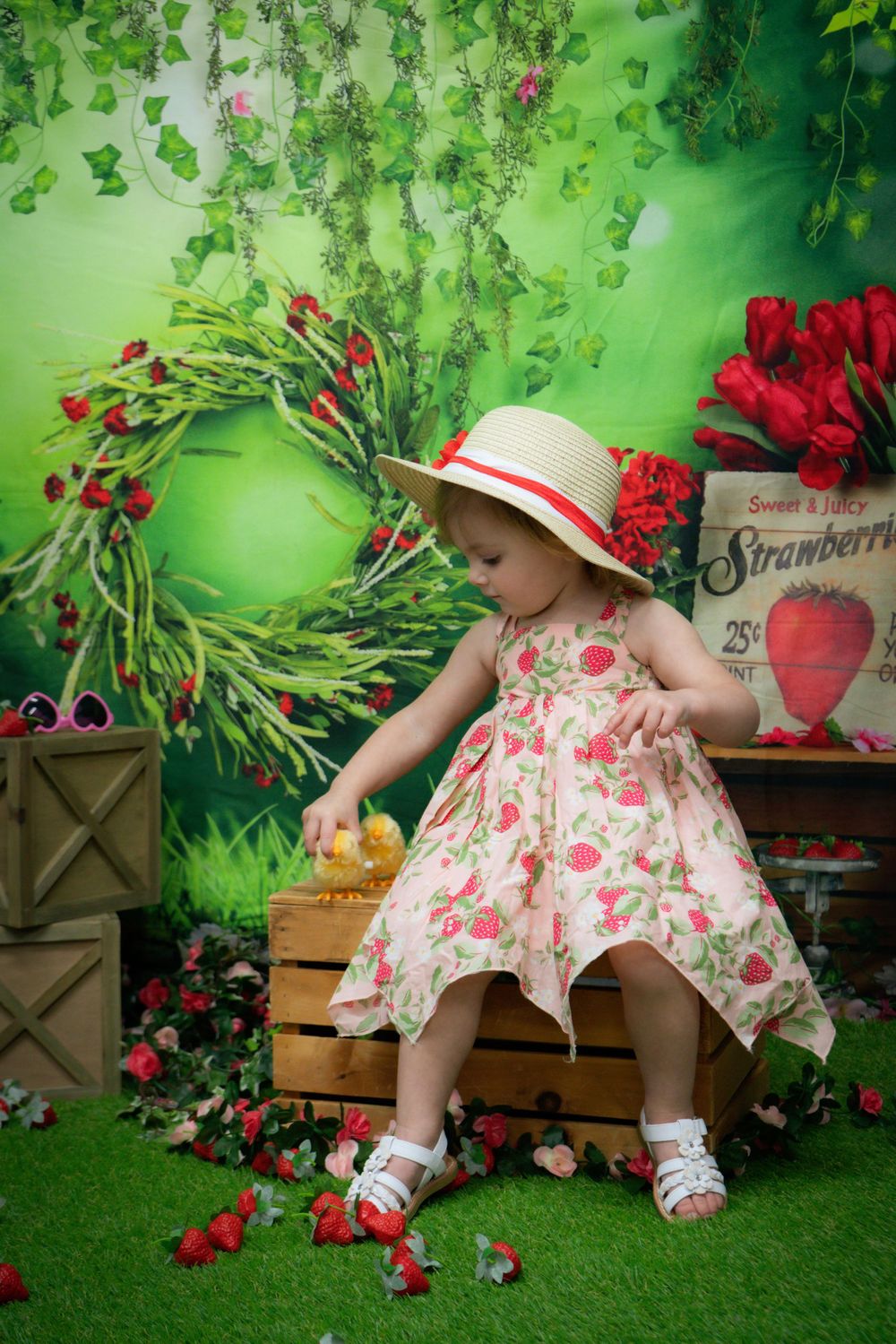 A toddler in a floral dress and sun hat plays in a garden setting with strawberry decorations.
