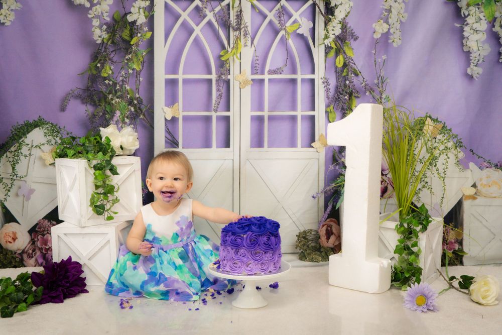 A baby in a blue dress sits among floral decorations against a purple backdrop with white arched windows.