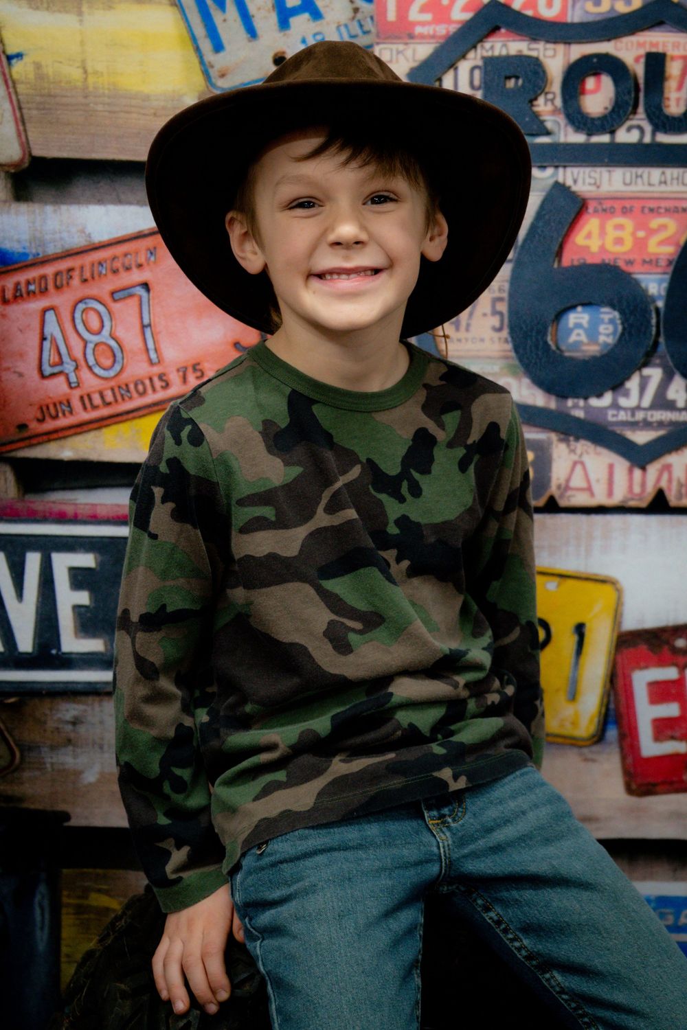 A child wearing a black cowboy hat and camouflage shirt sits against a backdrop of colorful vintage signs.