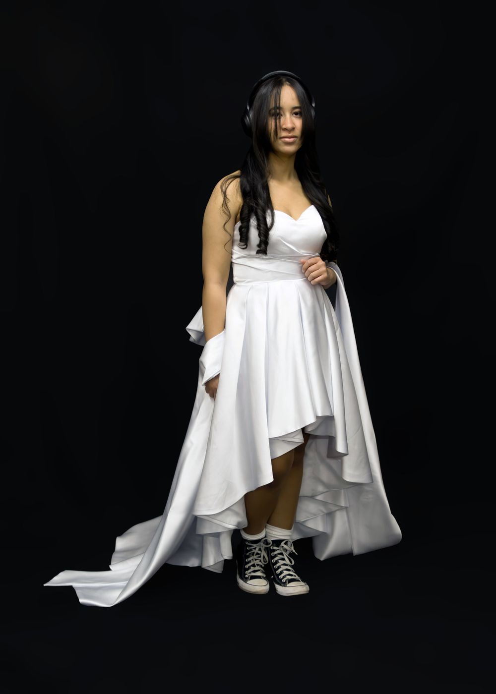 Person in white high-low dress and headphones poses against black background.