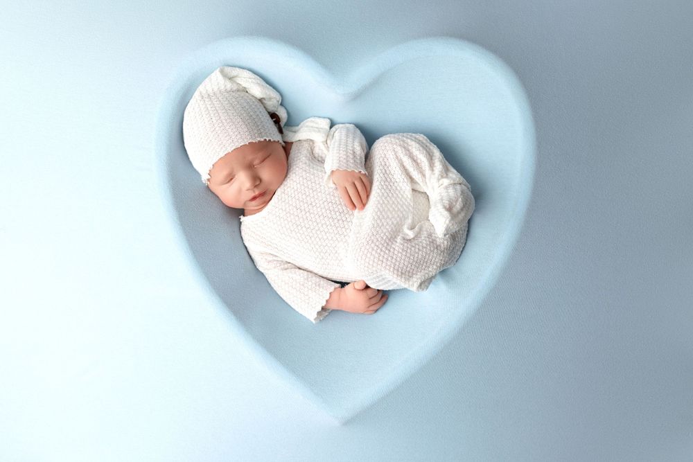 Sleeping newborn in white knit outfit laying in heart-shaped blue backdrop during studio session.