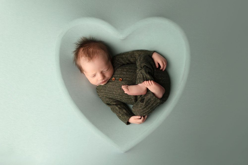 Sleeping newborn in cozy knit sweater photographed from above in heart-shaped frame on mint green background.