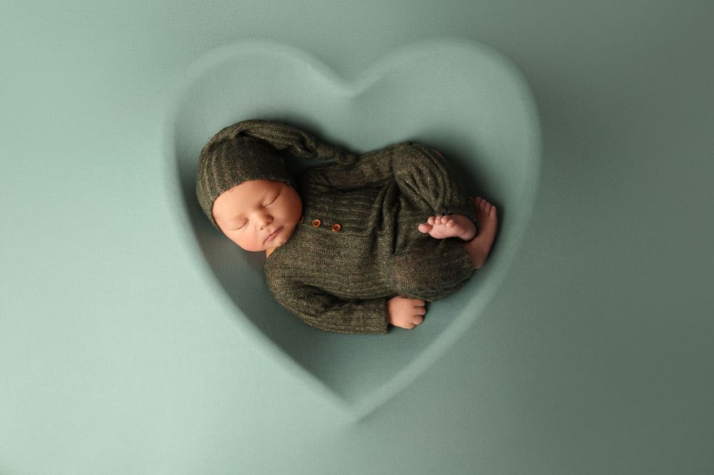 Sleeping newborn in brown knit outfit photographed in heart-shaped frame on mint background.