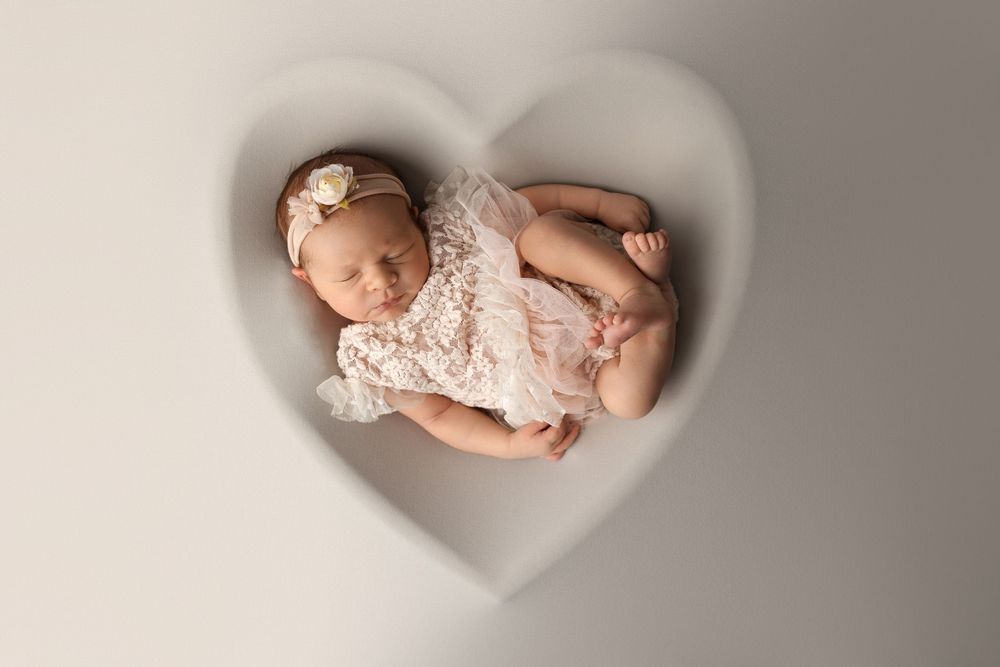 Sleeping newborn baby wearing white lace dress lying on soft beige backdrop in studio lighting.