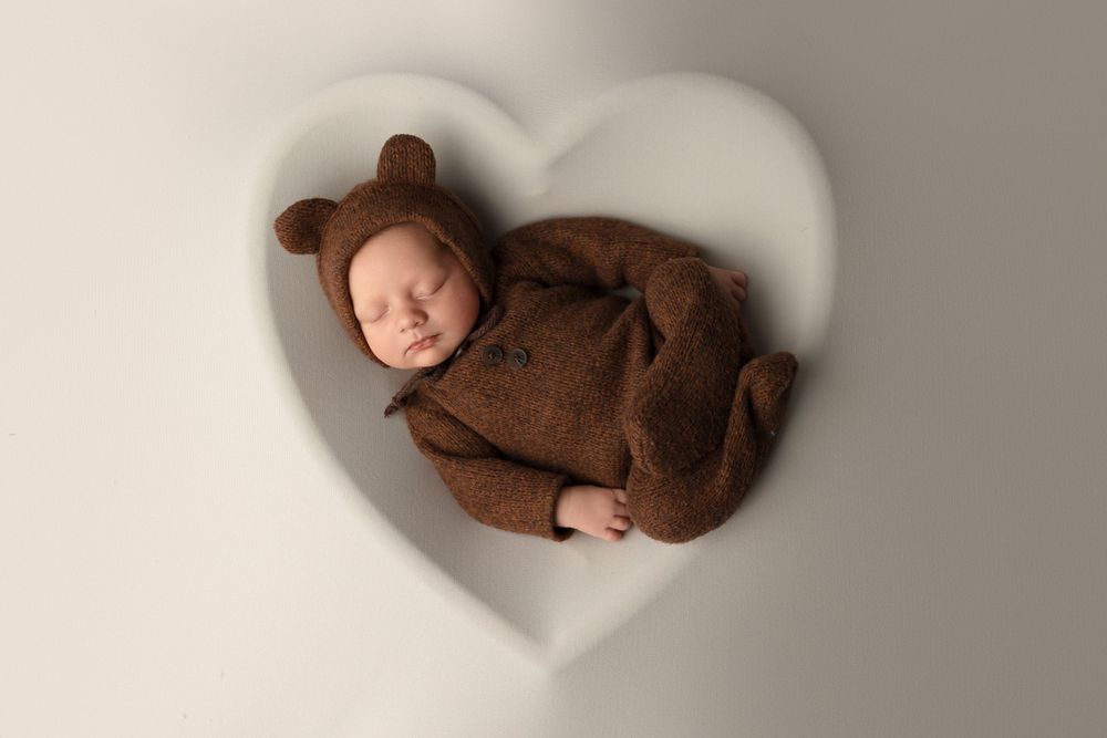 Baby wearing brown bear costume sleeping in heart-shaped prop on light background.
