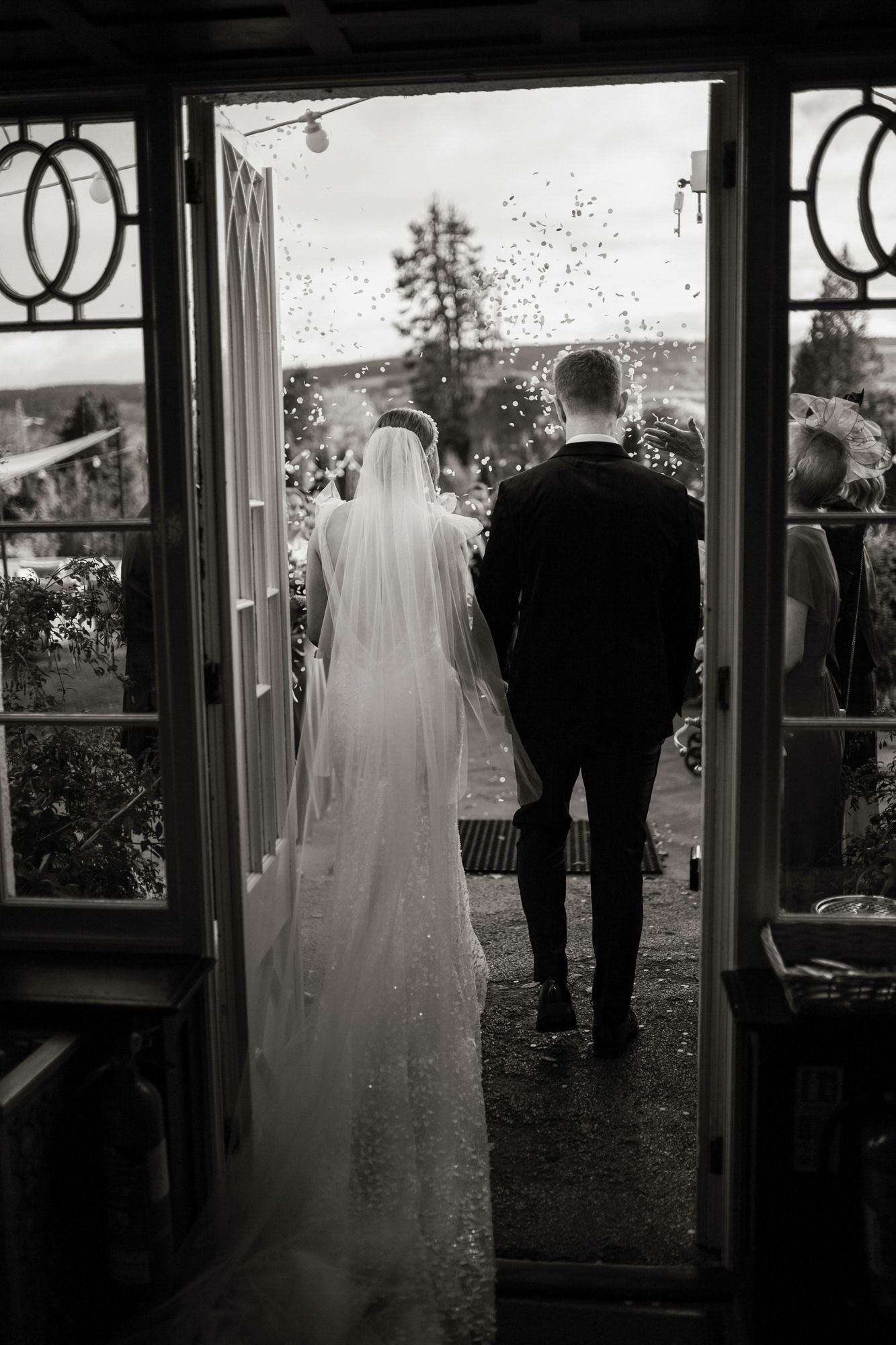 A black and white wedding photo showing a couple standing in a doorway with a flowing veil and scenic view beyond.