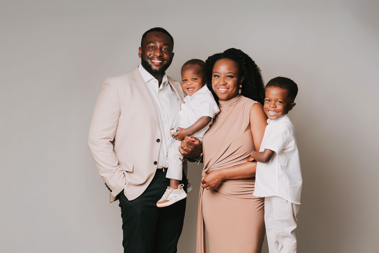 A joyful Black family of four poses together in a bright studio, wearing coordinated beige and white outfits.