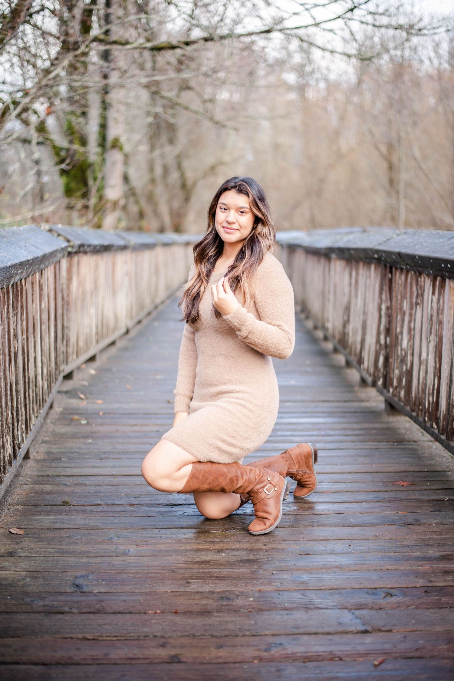 Person in beige dress and boots poses playfully on wooden boardwalk.