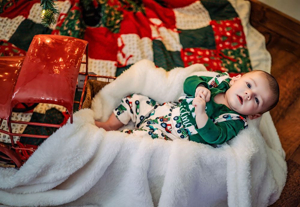 A baby in a white blanket and green outfit lies on the floor surrounded by wrapped Christmas presents and decorations.