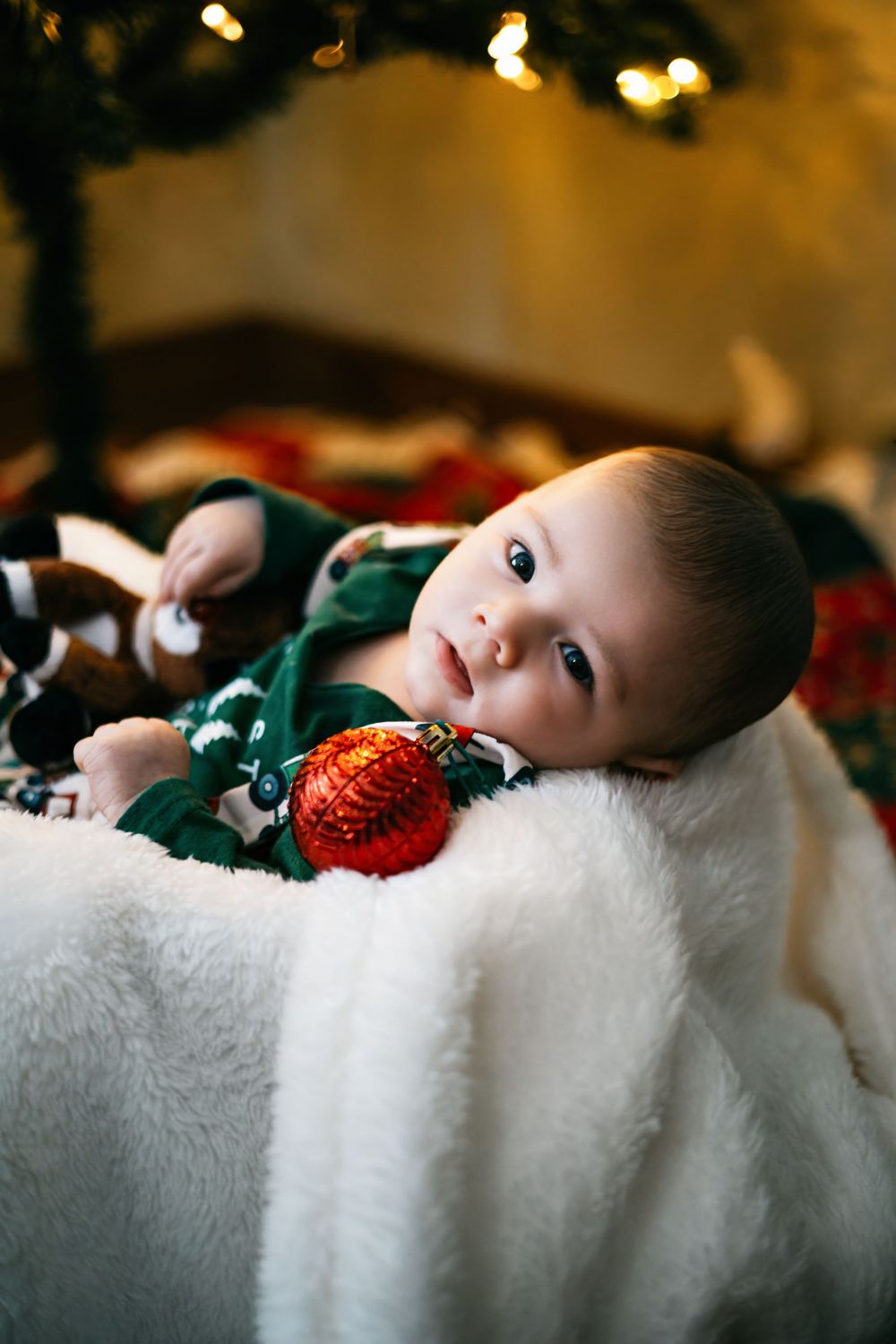 A baby lies on white fur with Christmas decorations and twinkling lights in the background.