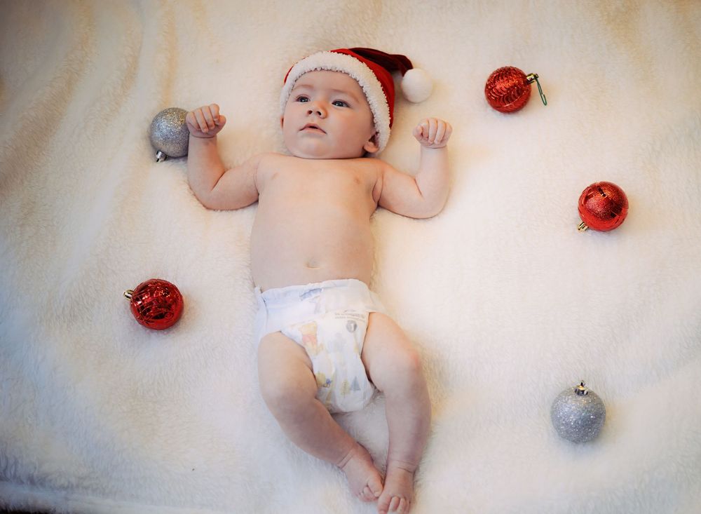 A baby lies on a white blanket surrounded by red and silver Christmas ornaments in a festive holiday photo.
