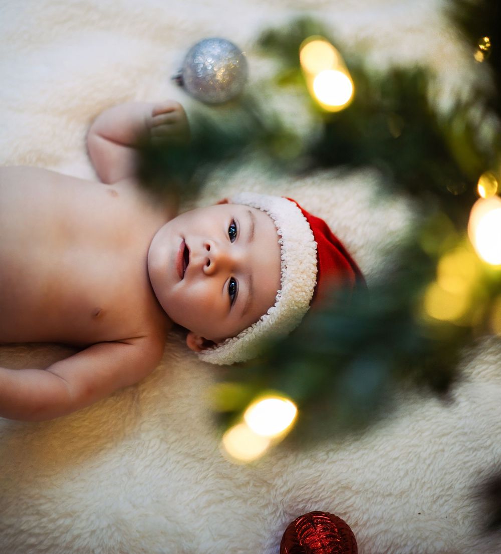 Close-up holiday photo of baby wearing Santa hat lying on white blanket with Christmas lights in background.