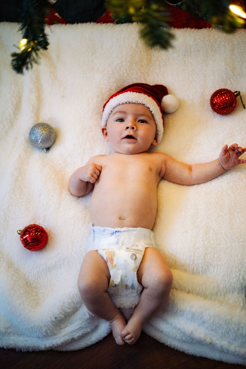 An infant wearing a Santa hat rests on soft white fabric surrounded by holiday ornaments.