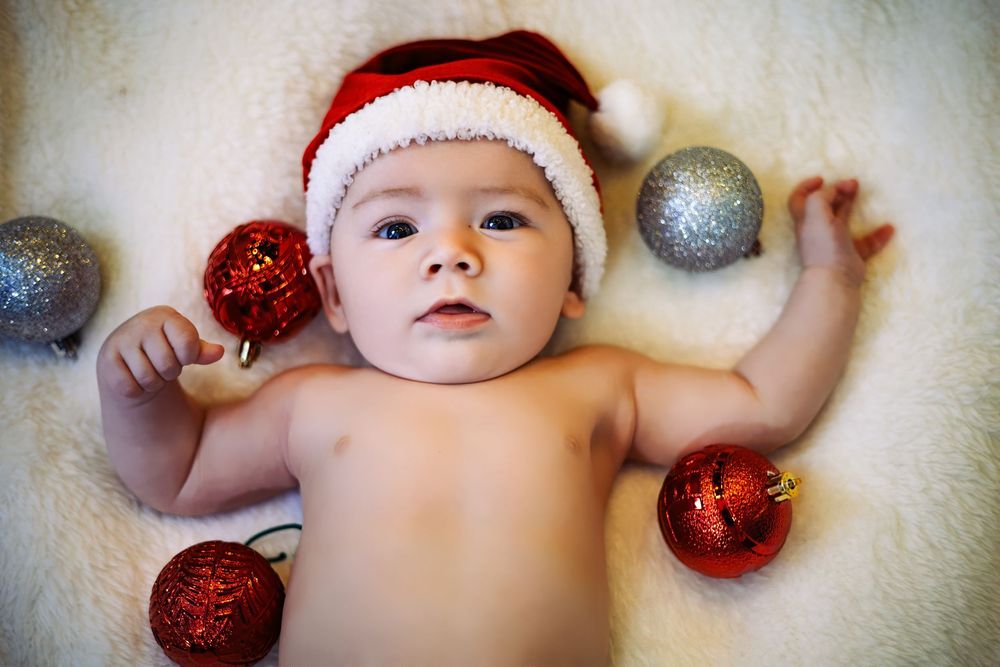 Baby wearing red Santa hat lying on white blanket with sparkly red and silver Christmas ornaments.