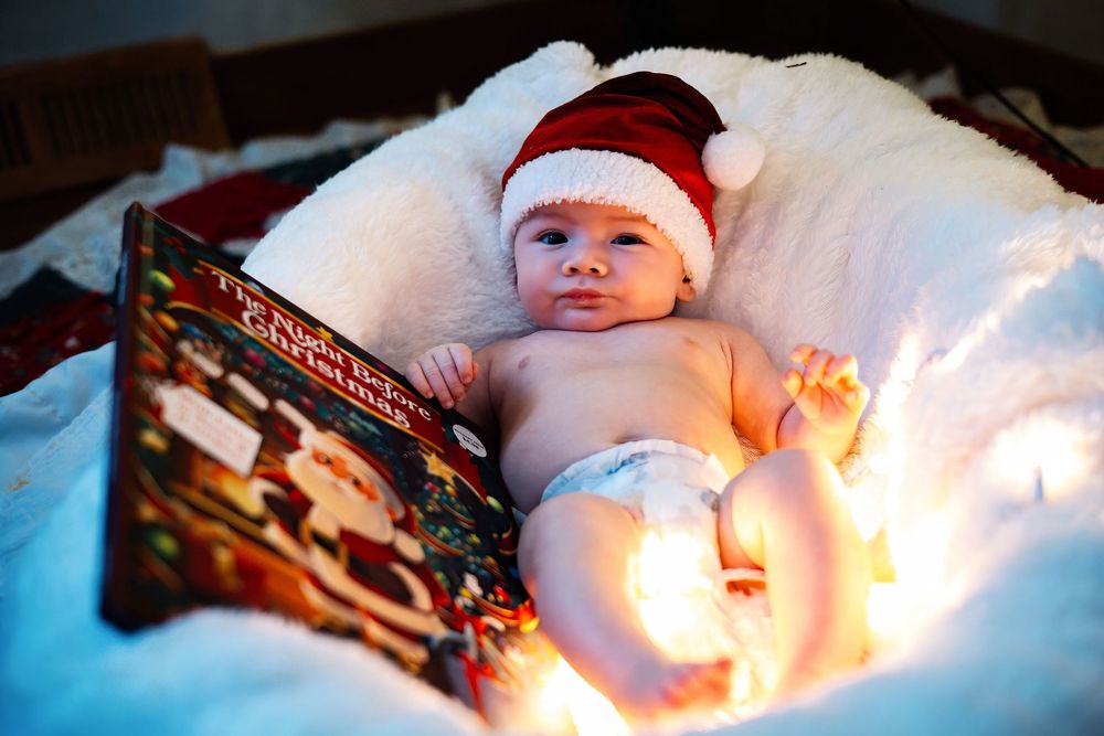Newborn baby wearing red Santa hat sleeps peacefully on white blanket during Christmas photo session.
