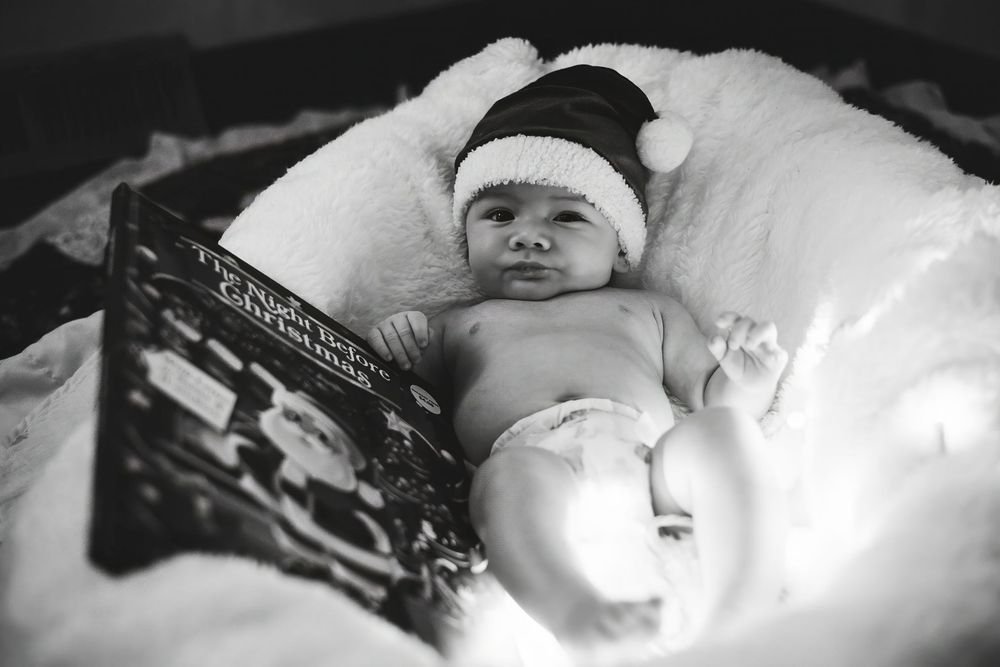 A black and white photo of a baby wearing a winter hat lying on white bedding.