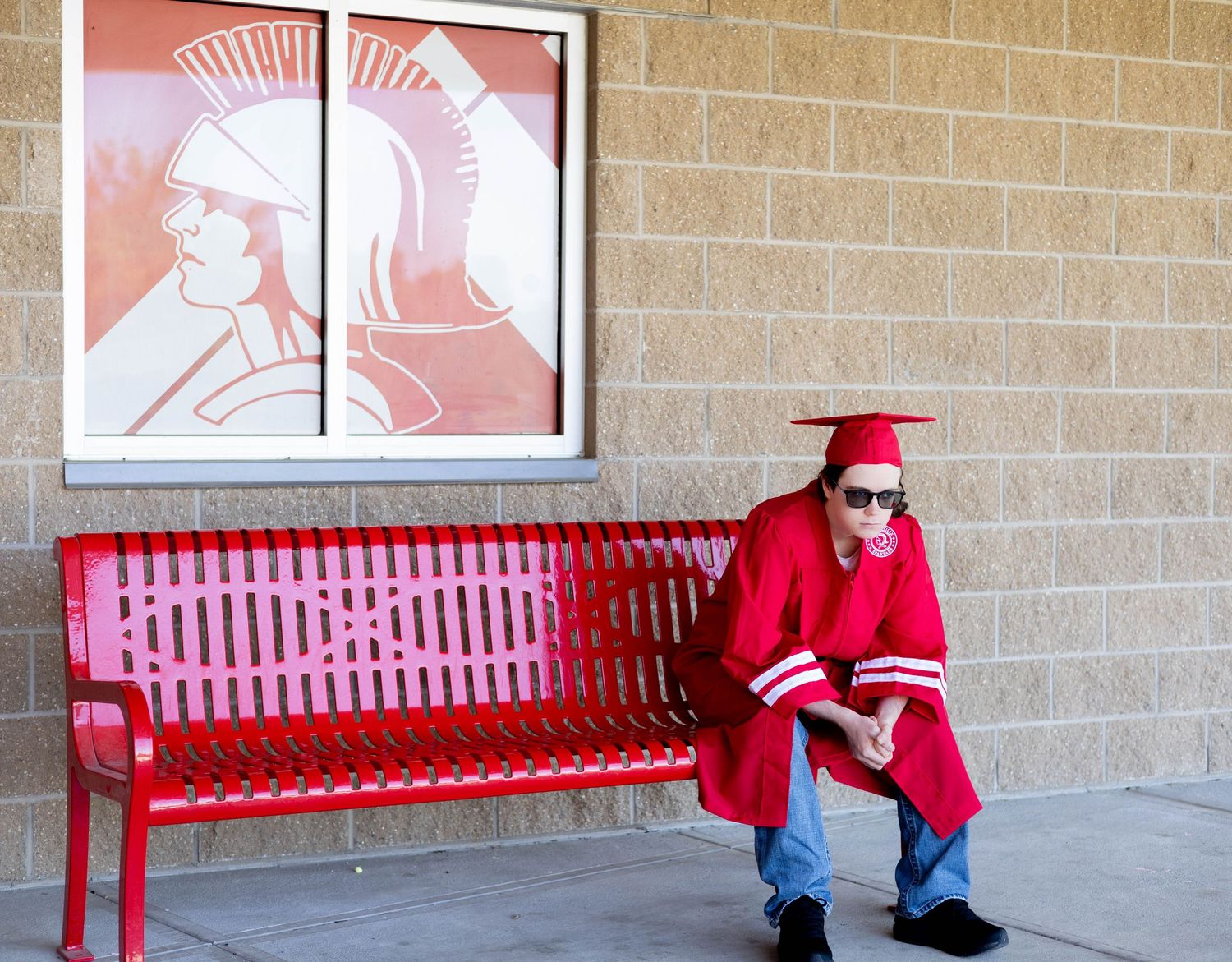 Graduate in red cap and gown sits reading on a red bench next to decorative artwork on brick wall.
