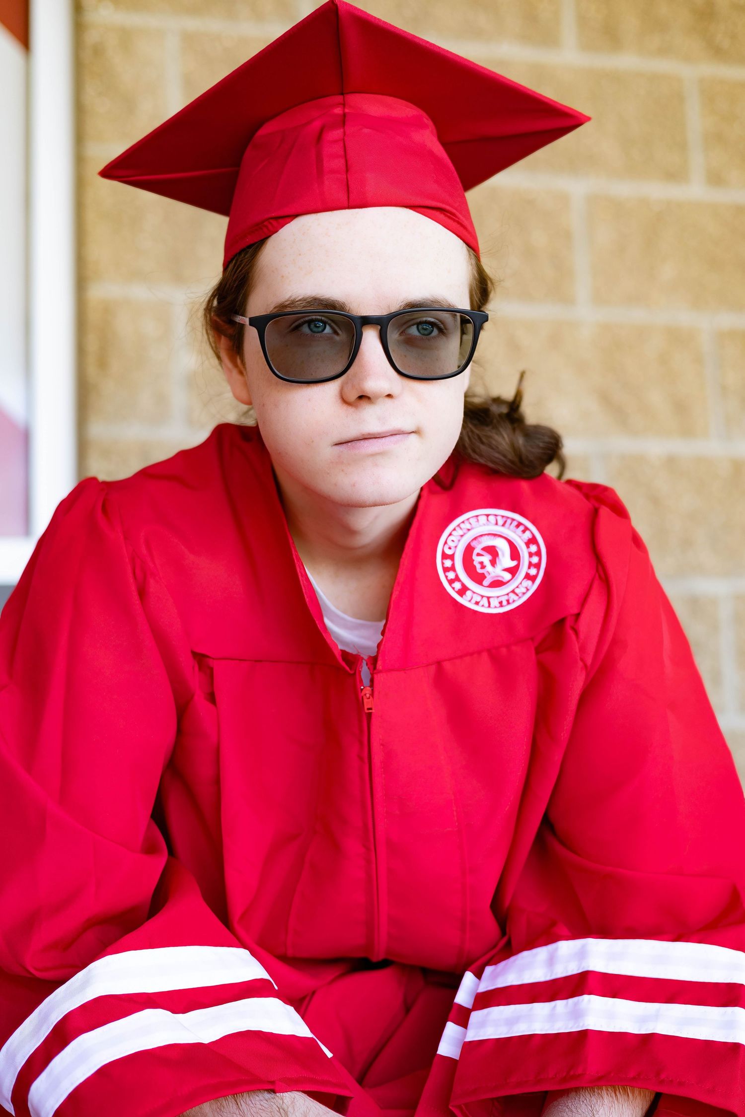 Graduate wearing red cap and gown with sunglasses poses against brick wall.