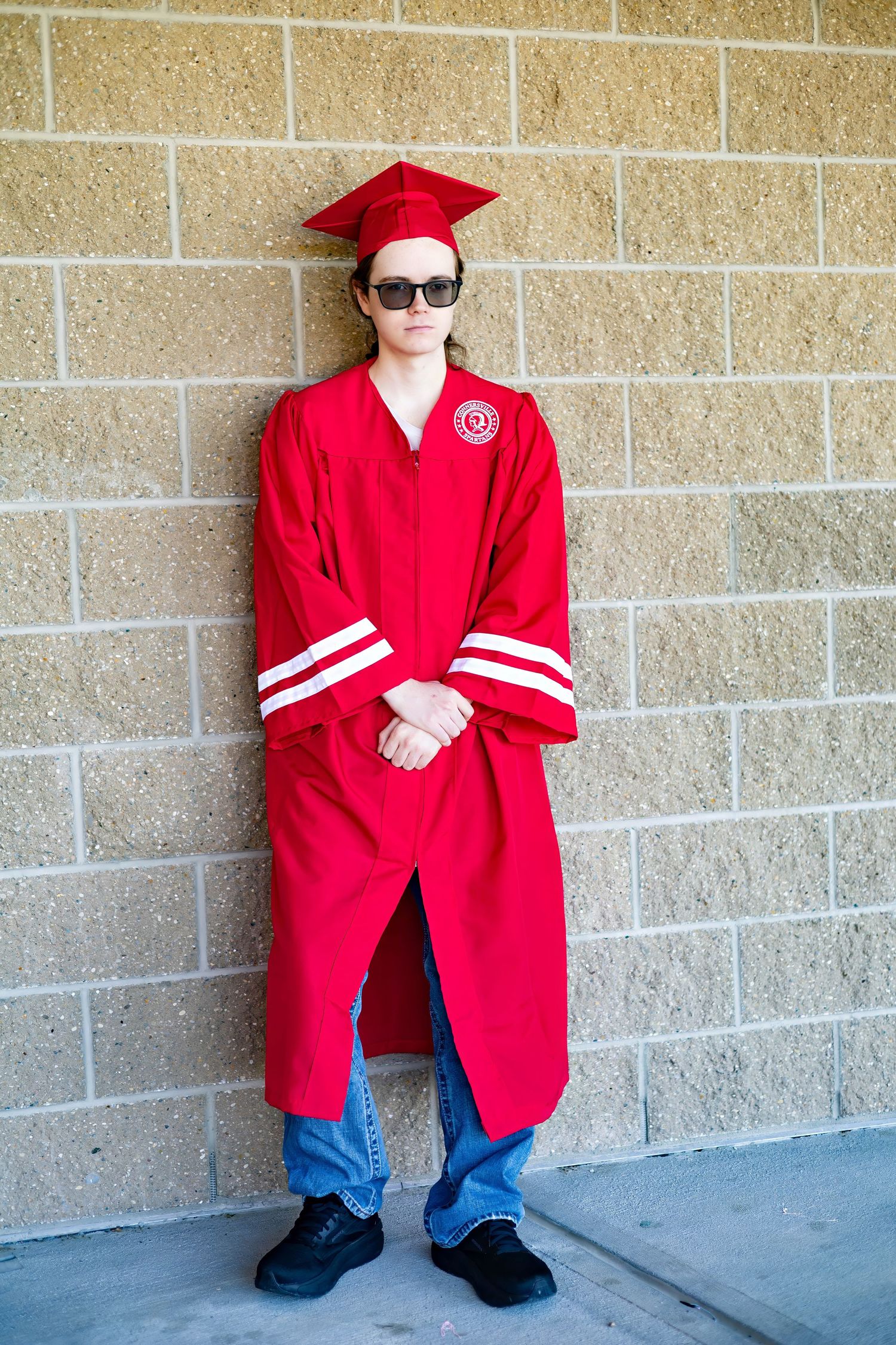 Graduate wearing red regalia and sunglasses stands against brick wall during commencement.