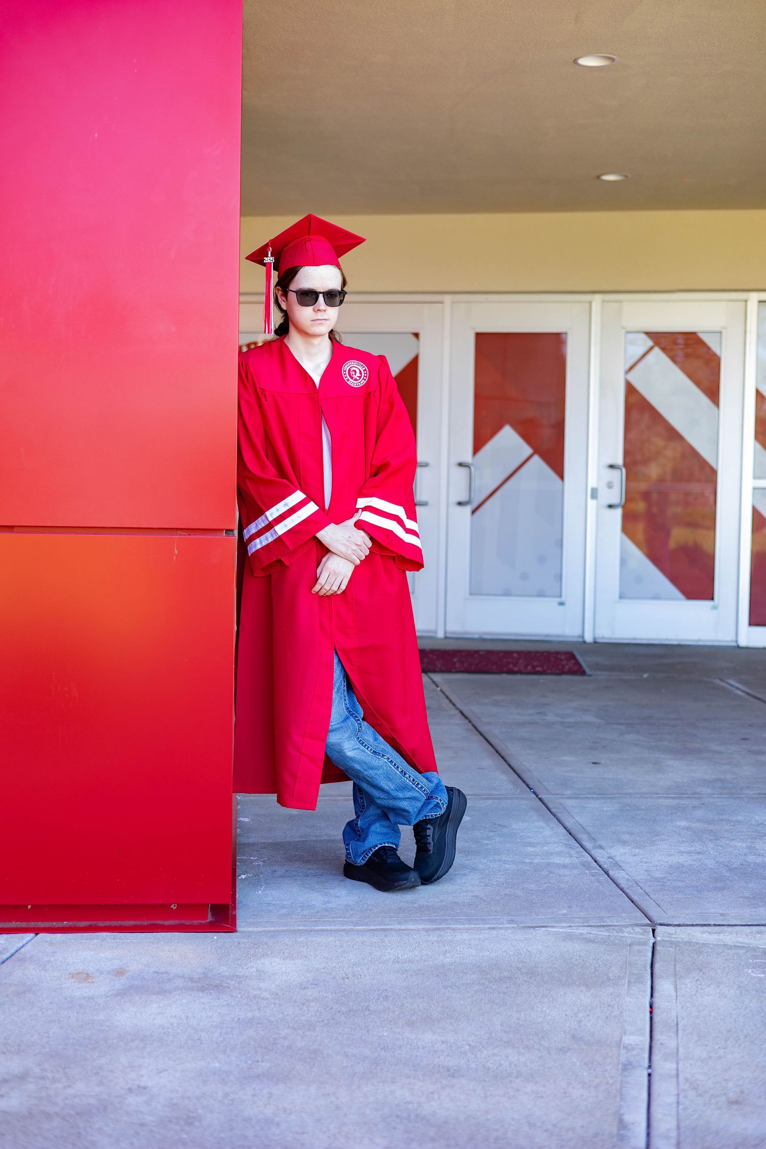 Graduate poses in red cap and gown against a bright pink wall and glass doors.