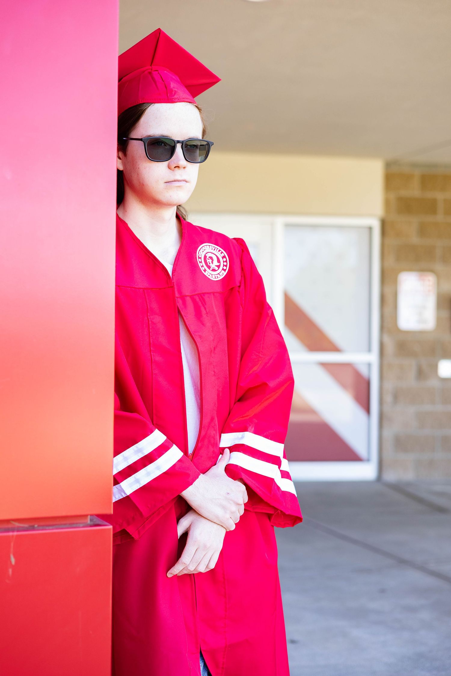 Graduate in red cap and gown with sunglasses poses confidently against a bright pink wall.