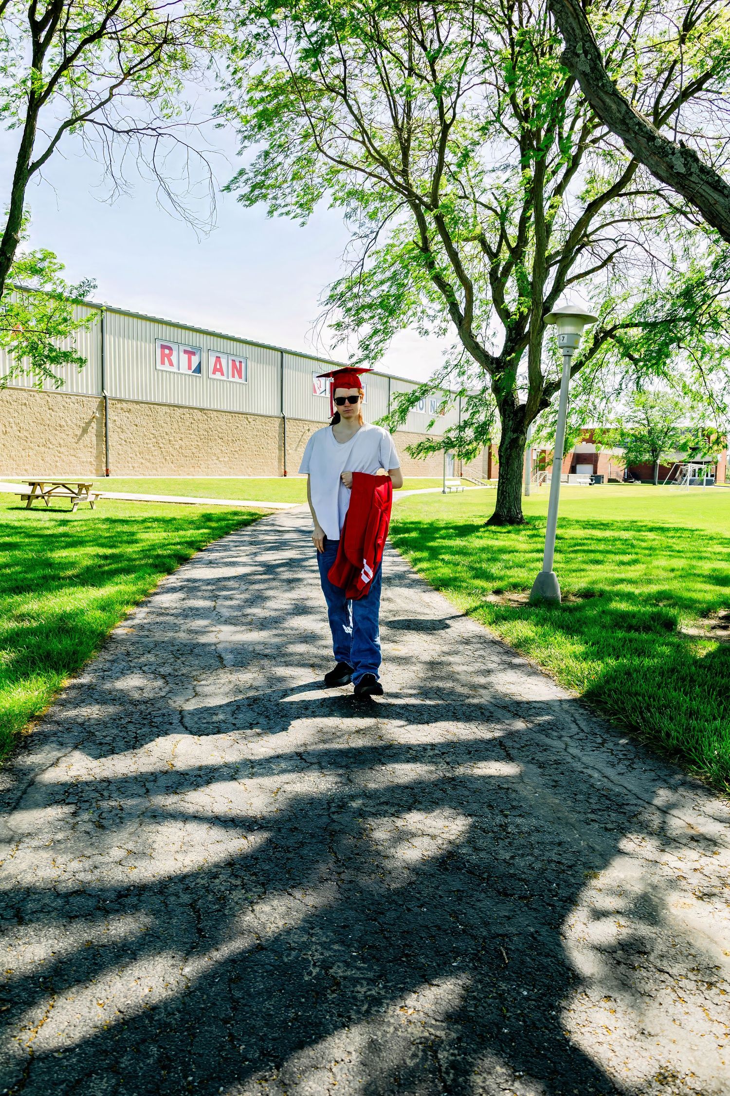 Graduate in red cap stands on pathway with school building in background.