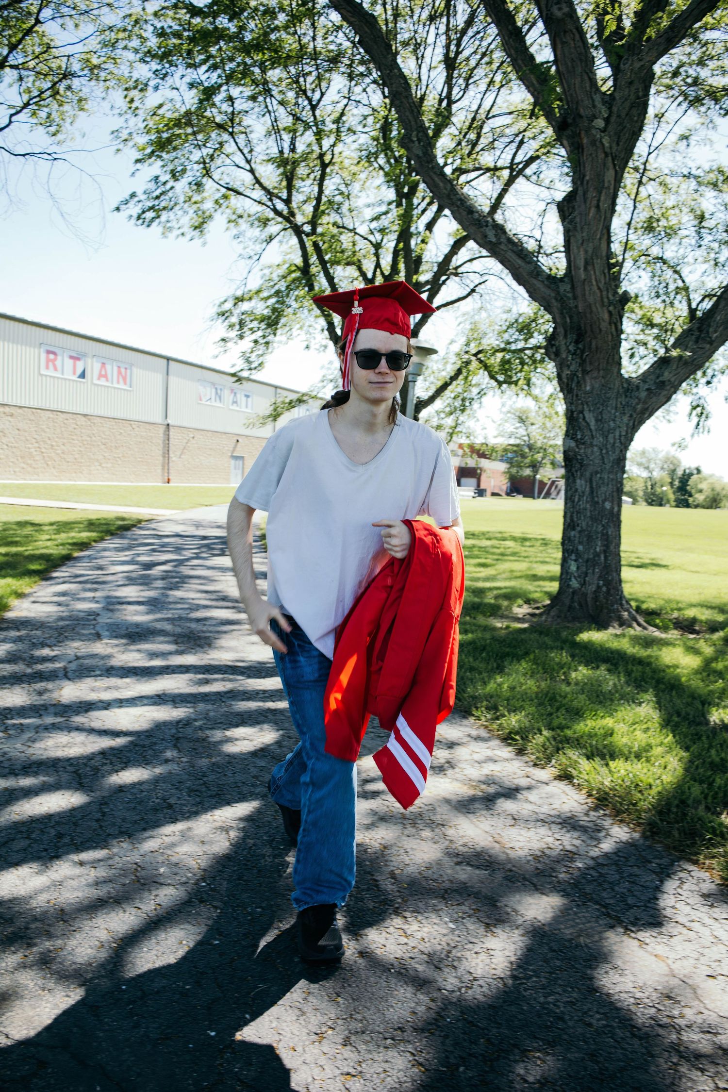 A graduate in casual attire holds red graduation robes while standing on a tree-lined path.