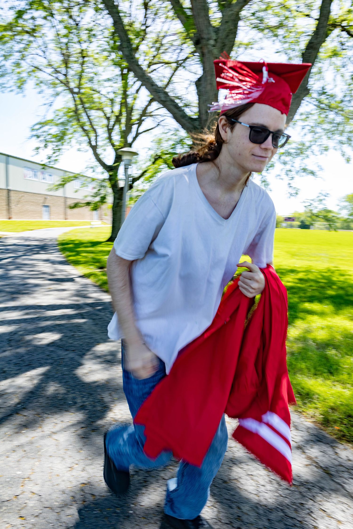 Graduate in casual attire and red cap walks outdoors carrying graduation gown on sunny day.