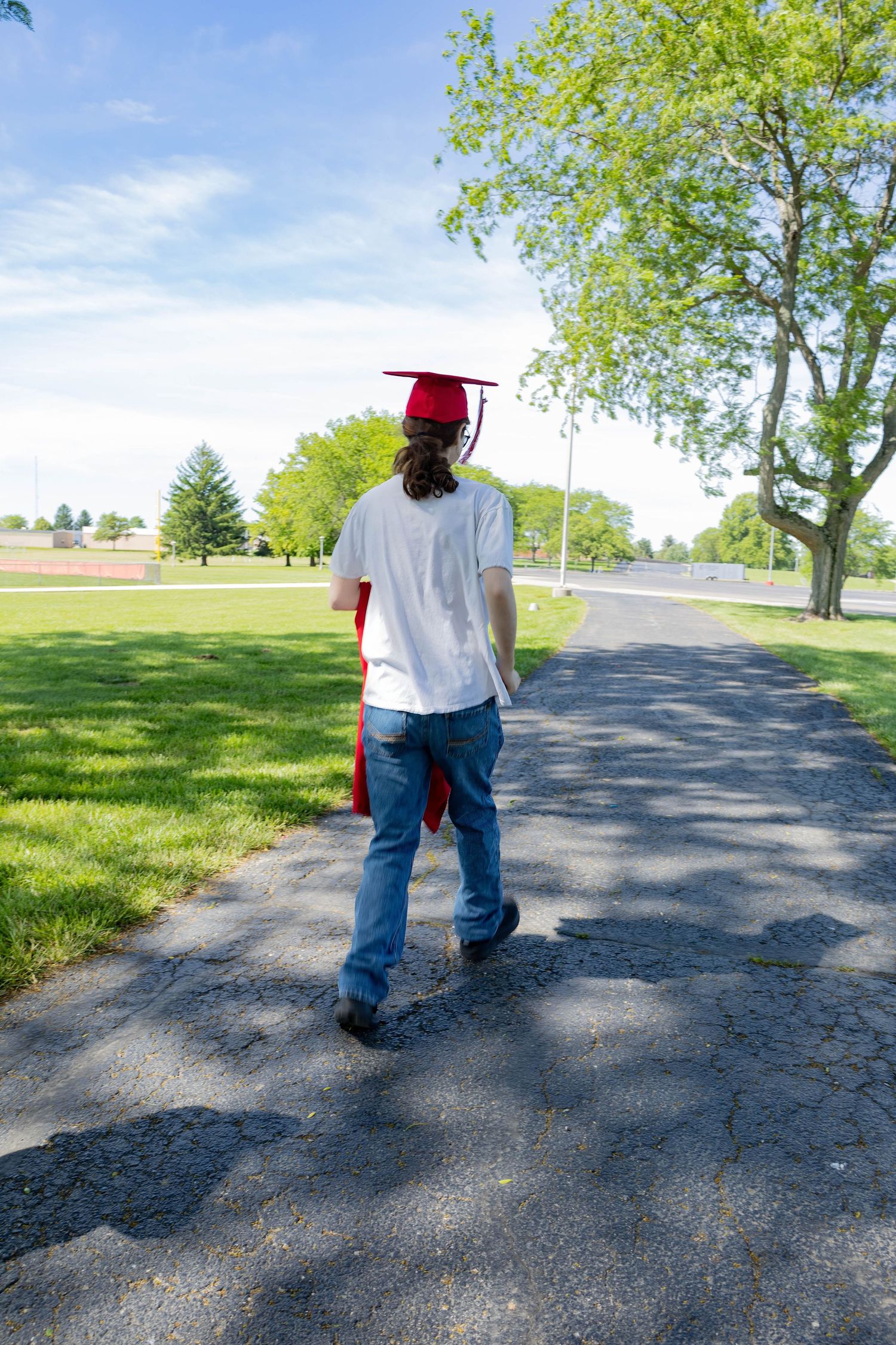 Graduate walks along shaded path carrying red graduation gown on sunny spring day.