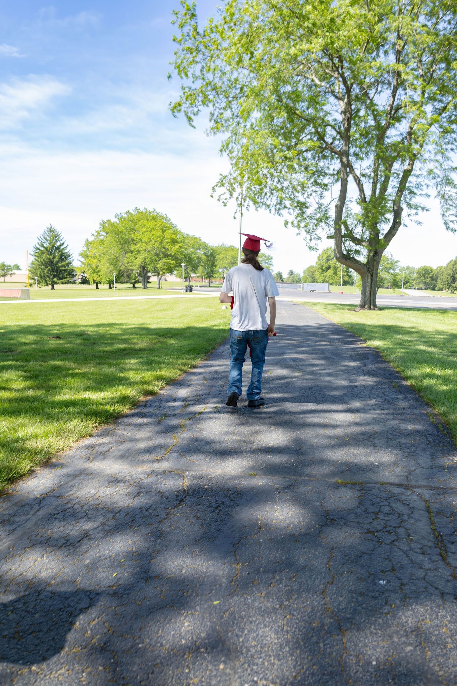 A graduate walks down a tree-lined path on a sunny day wearing casual clothes and a red cap.