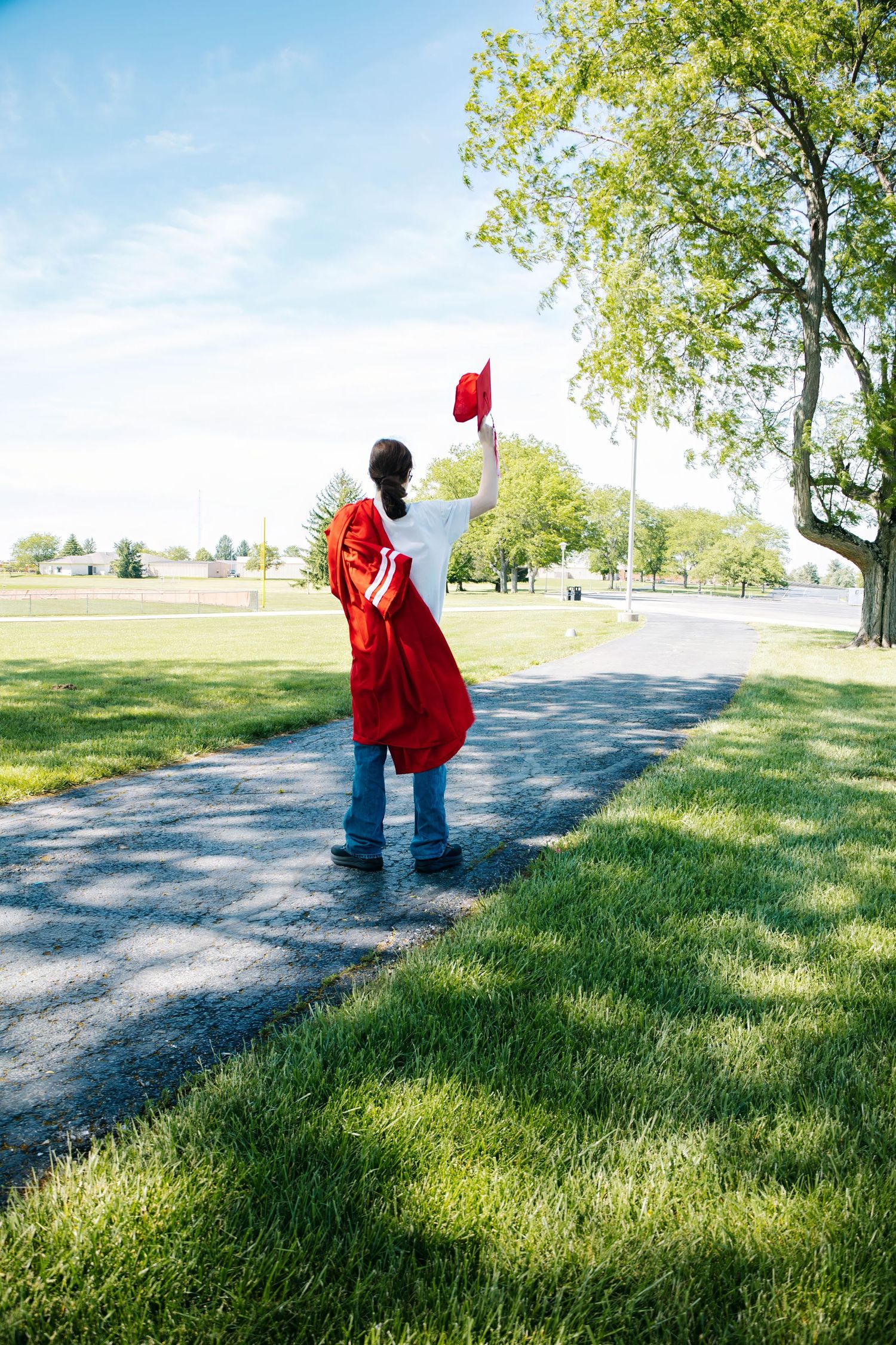 Graduate in red graduation gown raises cap triumphantly on a path surrounded by green grass and trees.