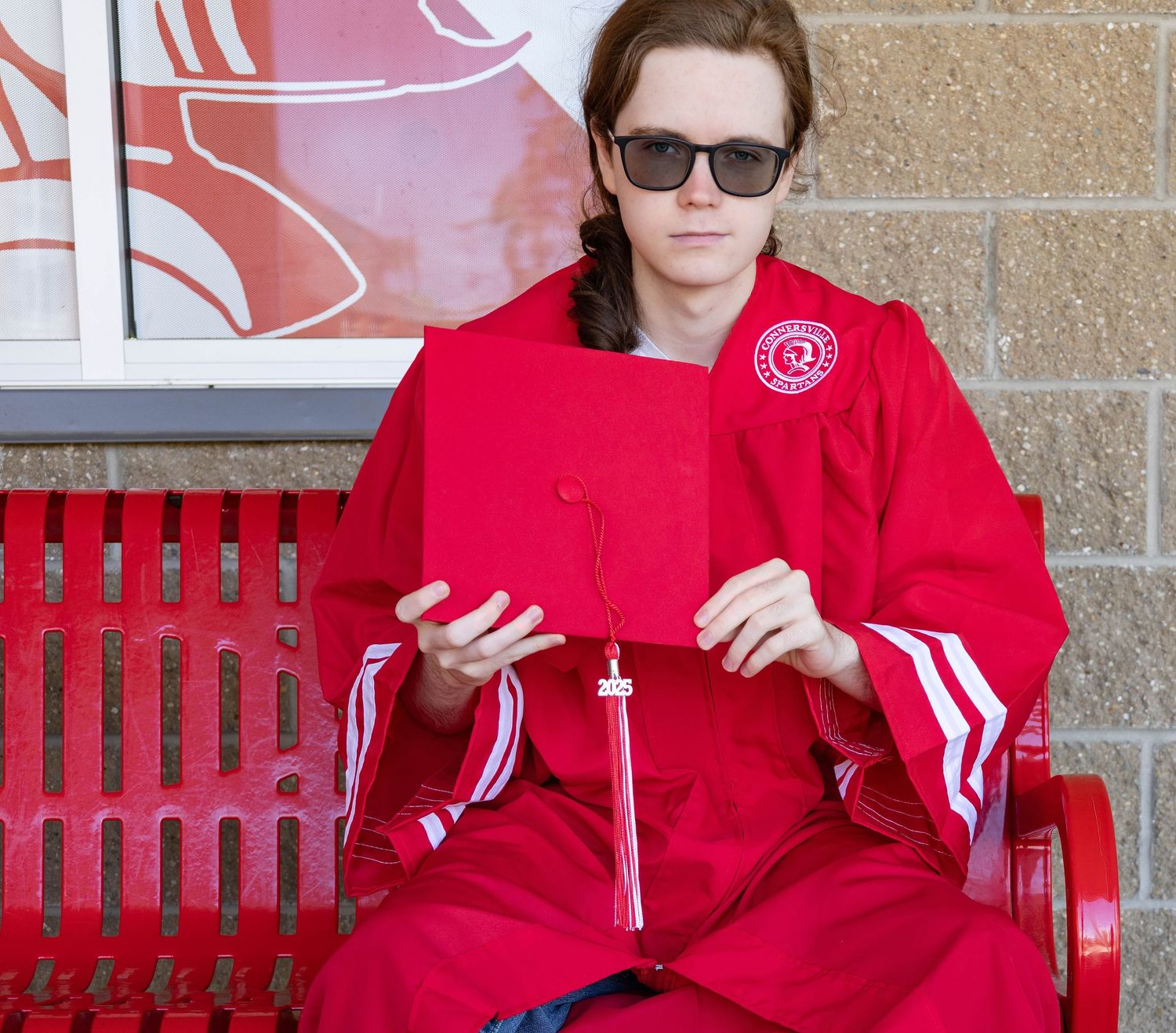 A graduate in red commencement robes sits on a red metal bench against a brick wall.