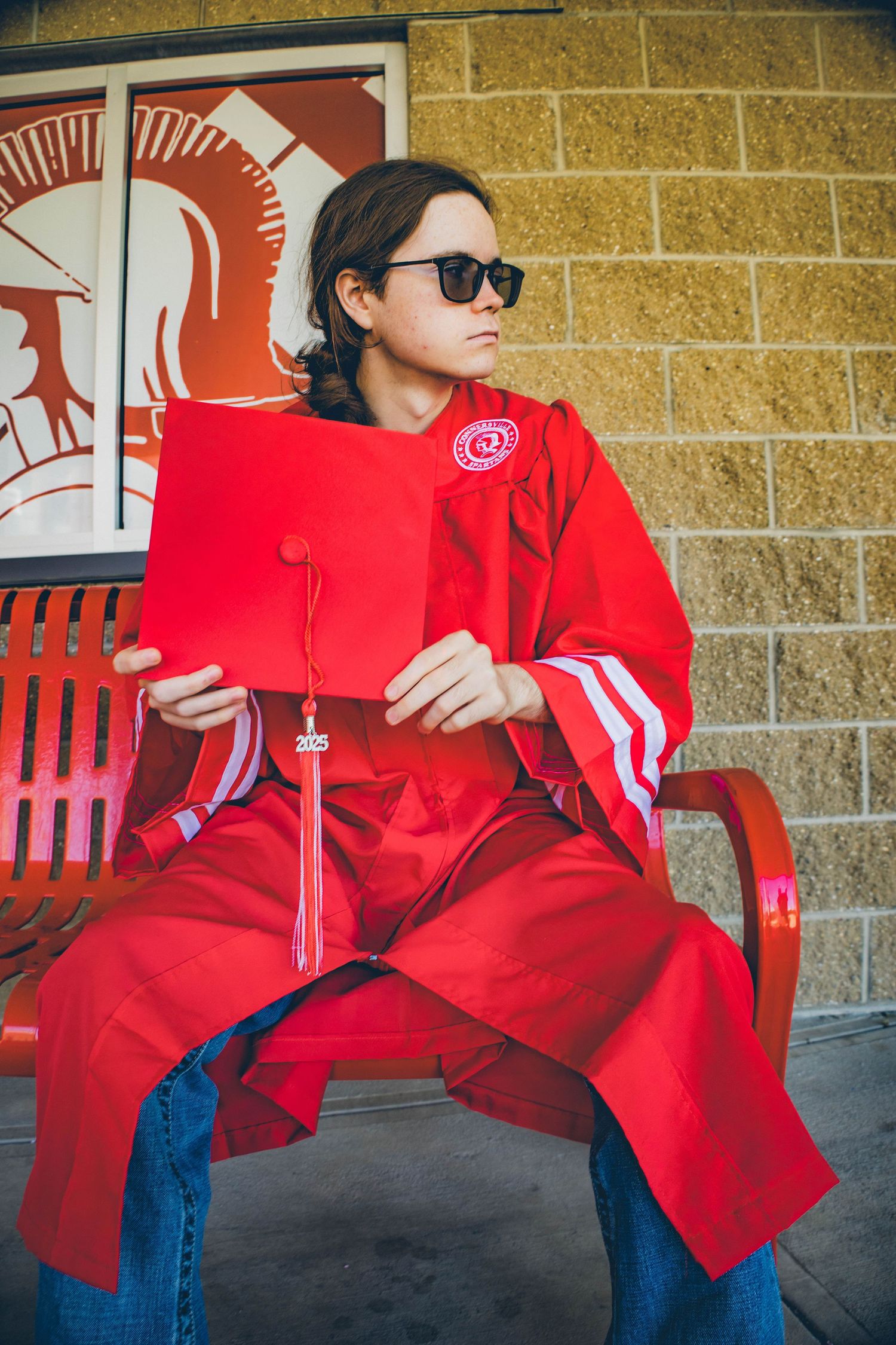 A graduate in red commencement robes holds their cap while sitting on a red bench.