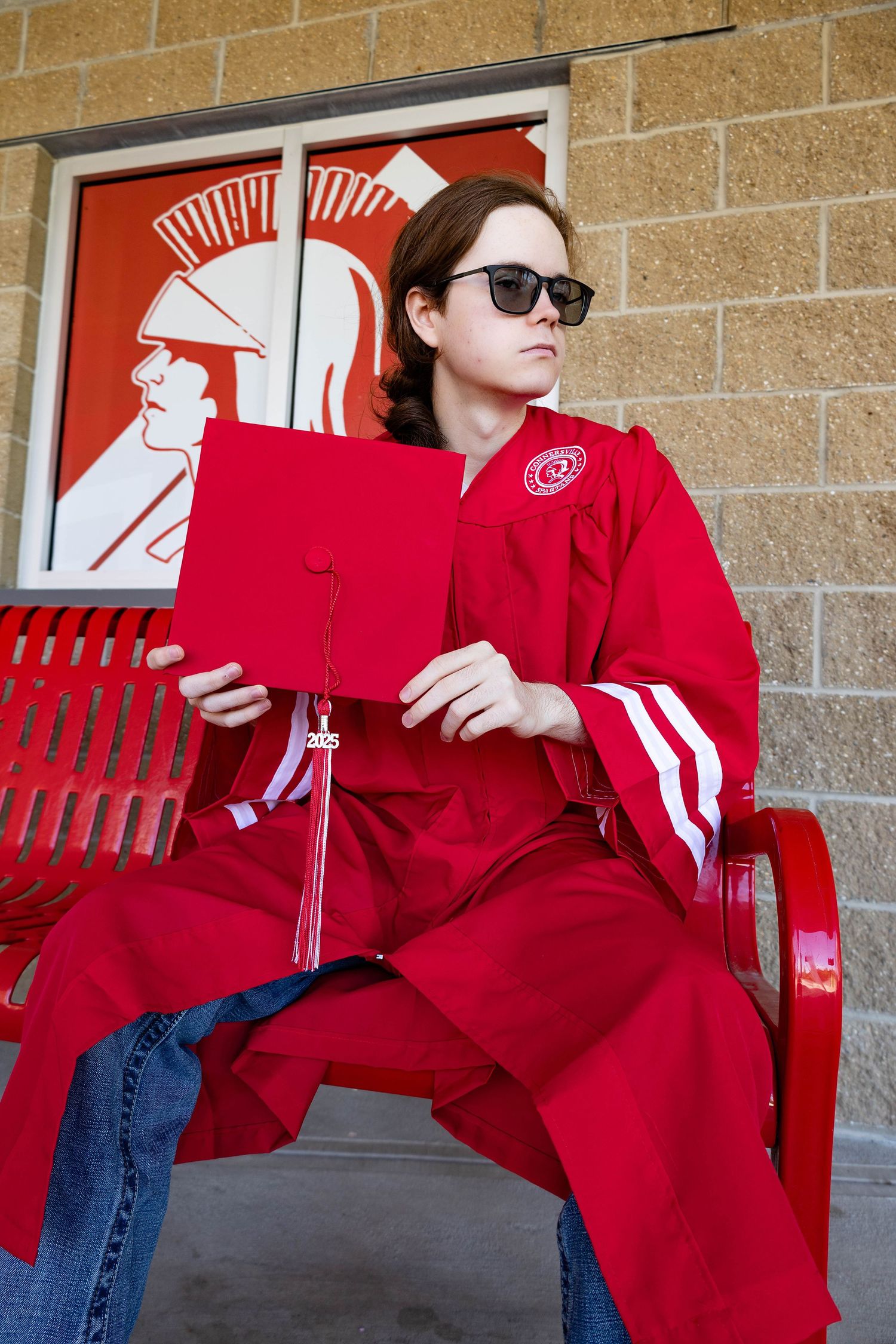 Graduate in red regalia and sunglasses sits with diploma near mascot wall art.