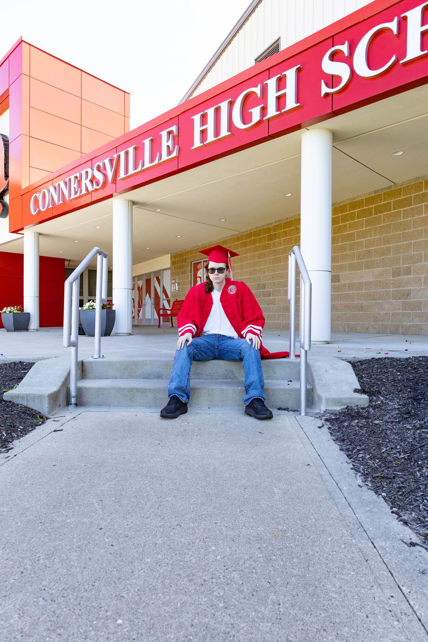 Graduate in bright red regalia sits on steps in front of Connersville High School entrance.