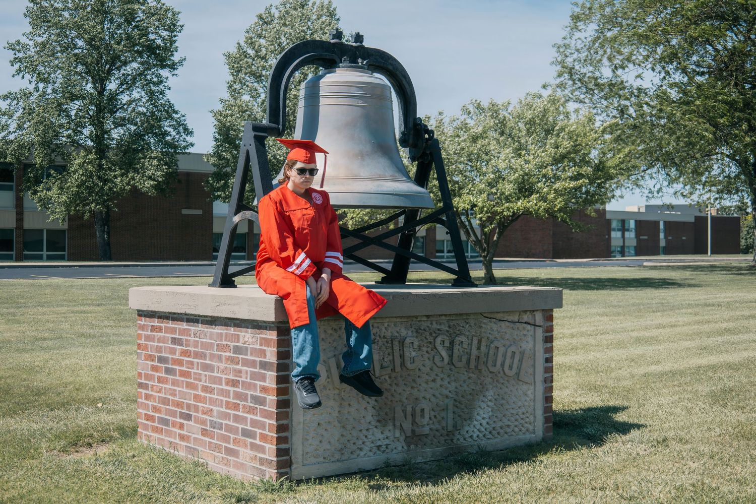 A graduate sits on a historic school bell monument wearing bright red graduation attire.