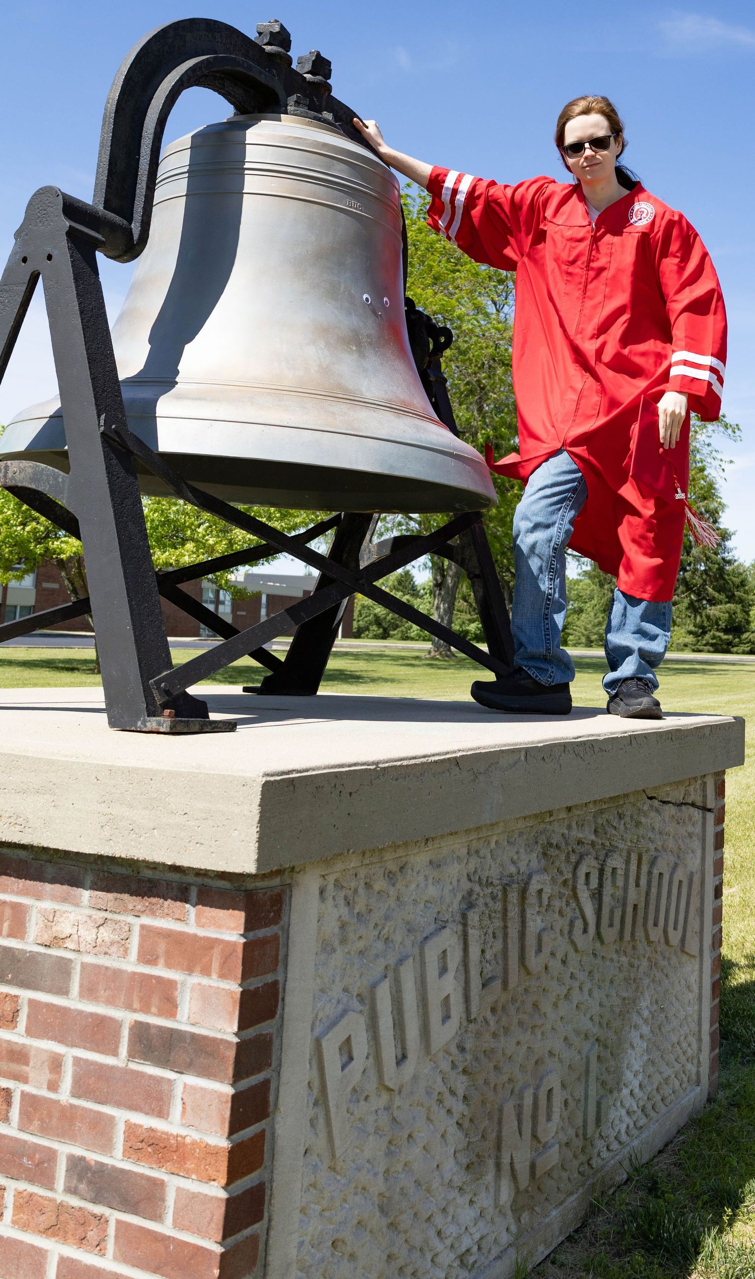 A person in red clothing poses next to a large historic bell mounted on a brick pedestal in a sunny outdoor setting.