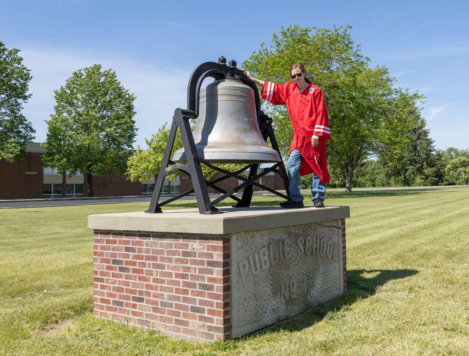 Graduate in red regalia poses with historic school bell mounted on brick pedestal.