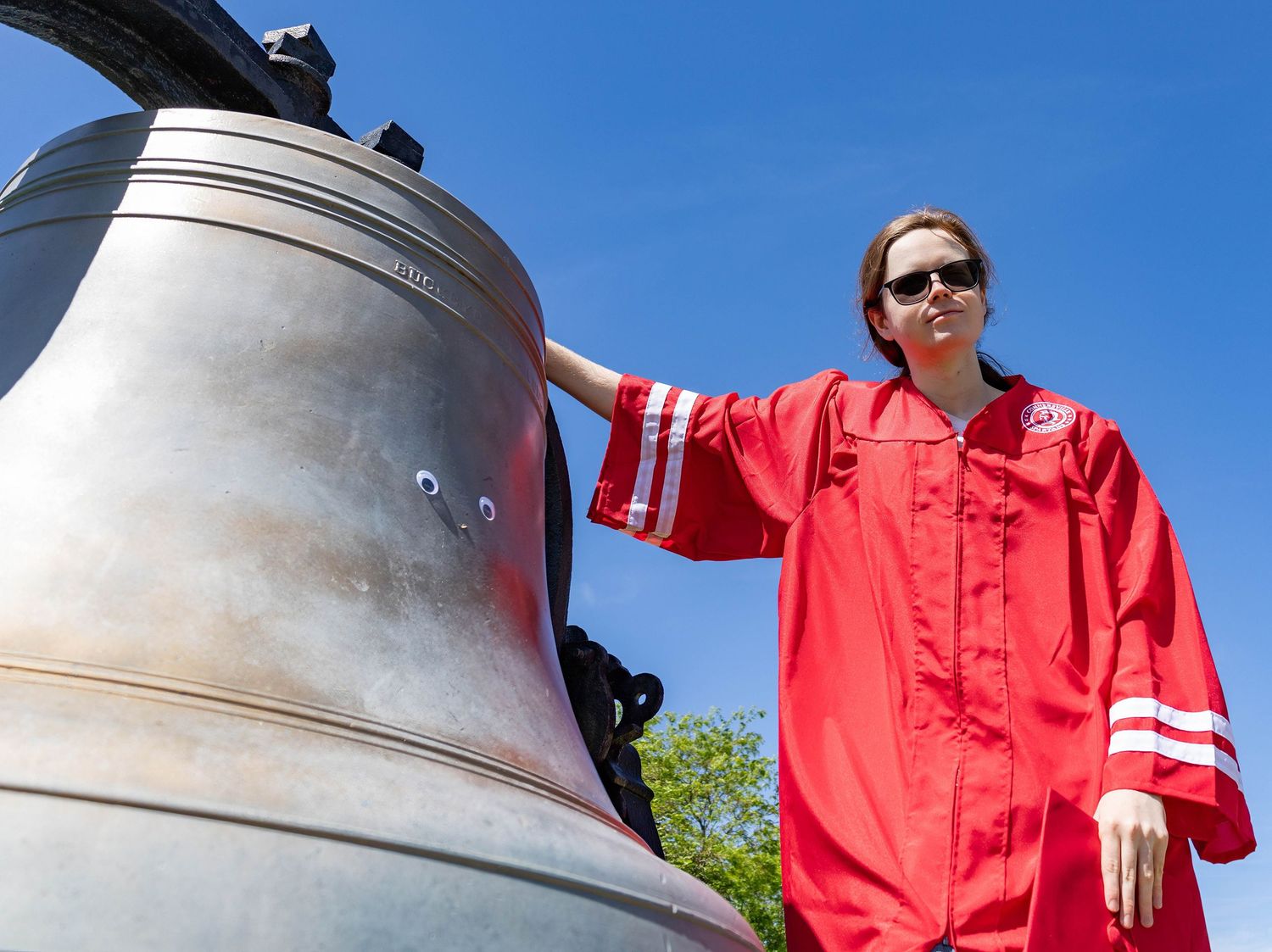 A graduate in red robes poses next to a large bronze bell against a bright blue sky.