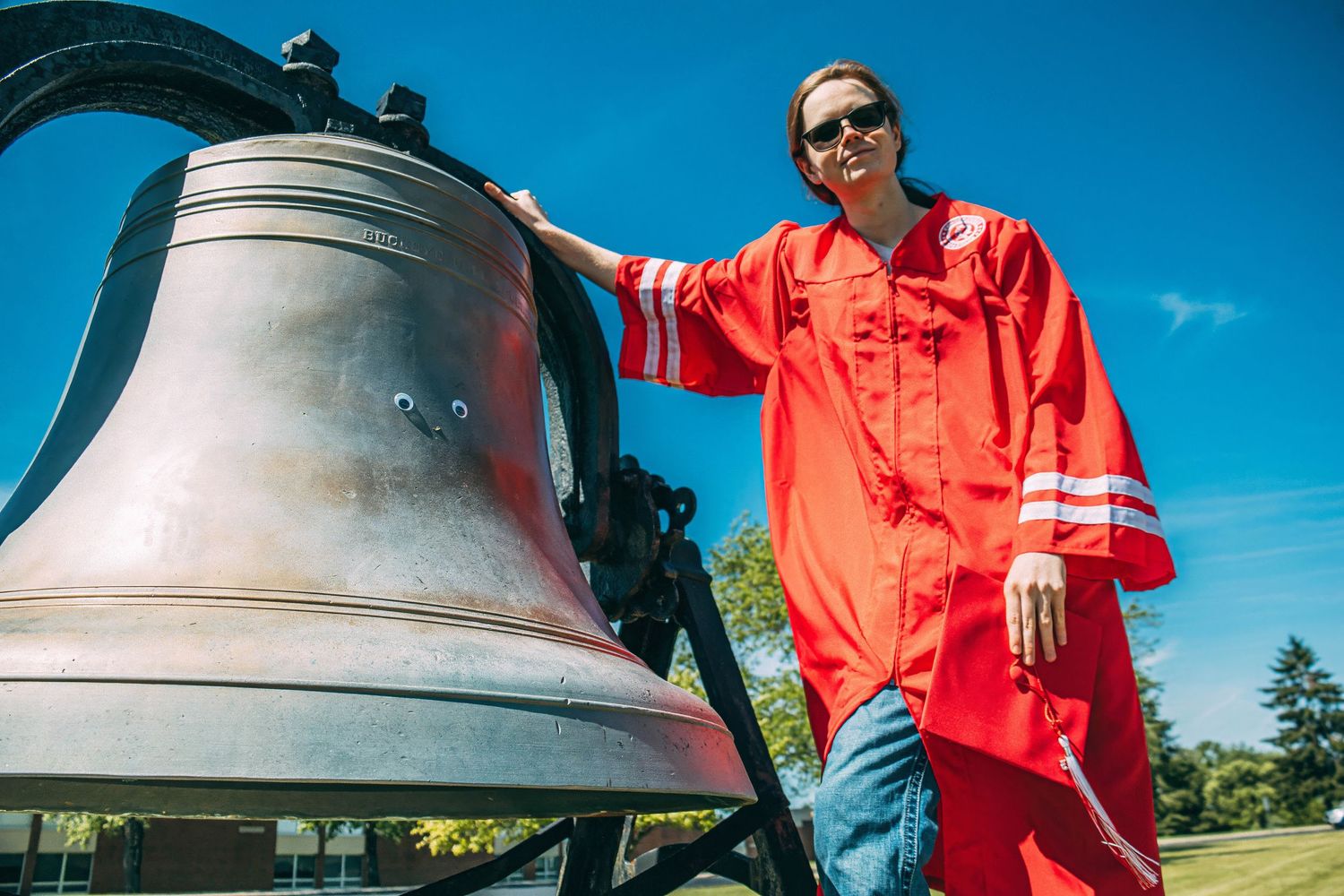Graduate in bright red gown poses with large antique bell against blue sky background.