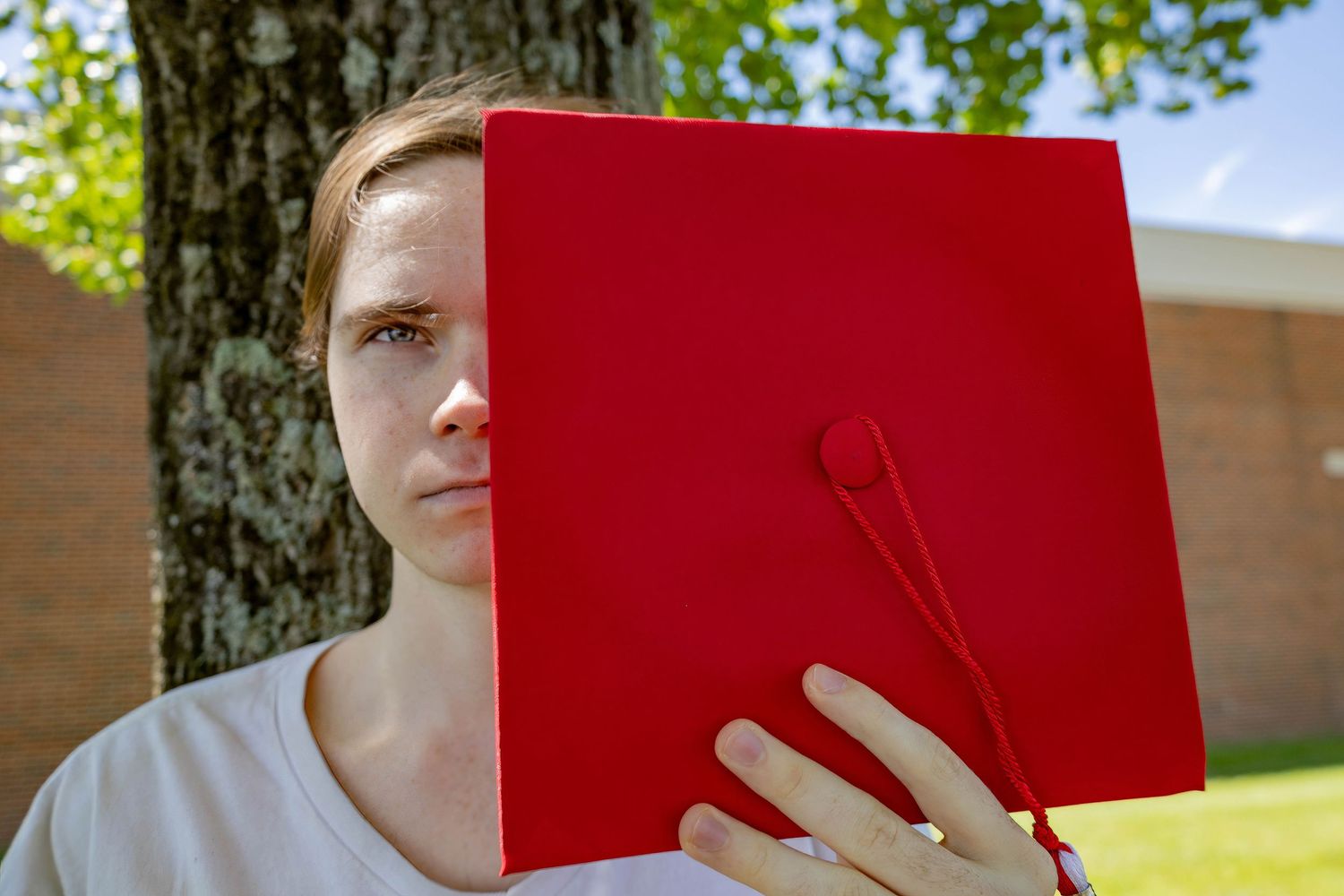 Graduate holding red diploma cover while standing behind tree.