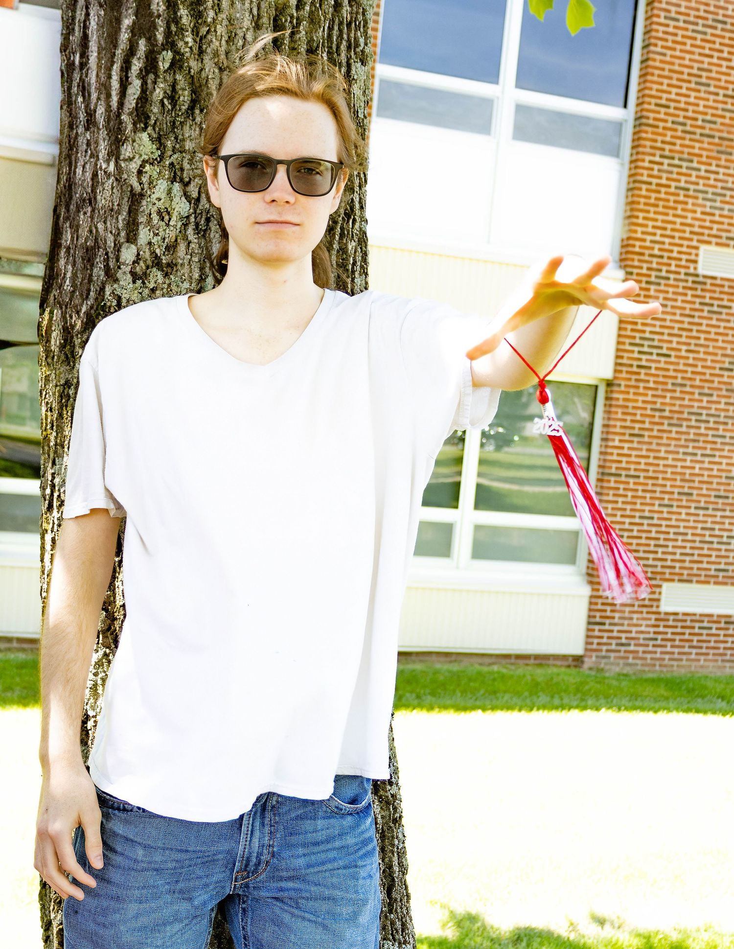 Person in white shirt and sunglasses leans against tree on school campus.