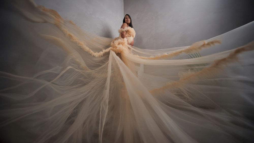 Ethereal portrait with billowing champagne fabric creating dramatic movement against dark backdrop.