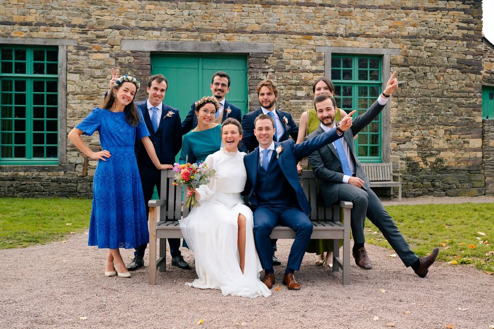 Wedding party poses playfully on rustic stone bench outside historic building with green windows.