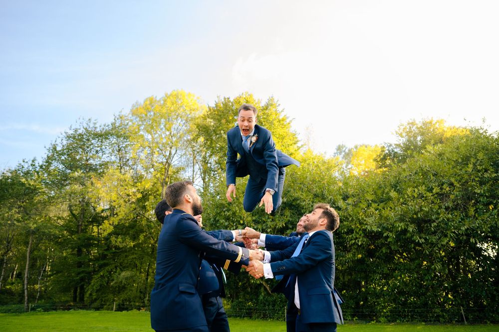 Two people in suits playfully fight in a garden setting with trees and bushes in the background.