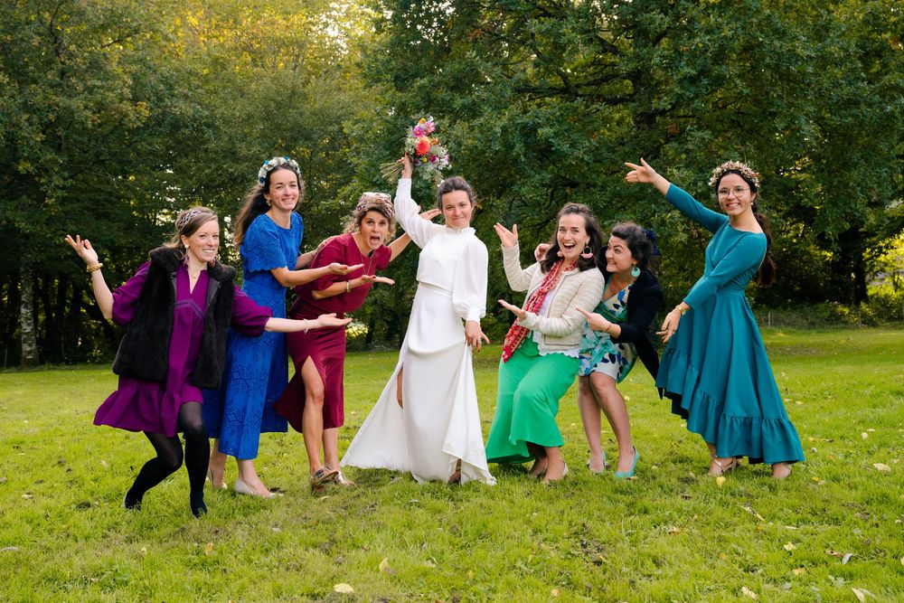 A group in colorful formal dresses strike playful poses together on a green lawn surrounded by autumn trees.