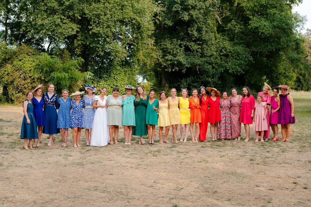 A colorful lineup of people wearing rainbow-ordered dresses stands together on a sandy beach for a group photo.