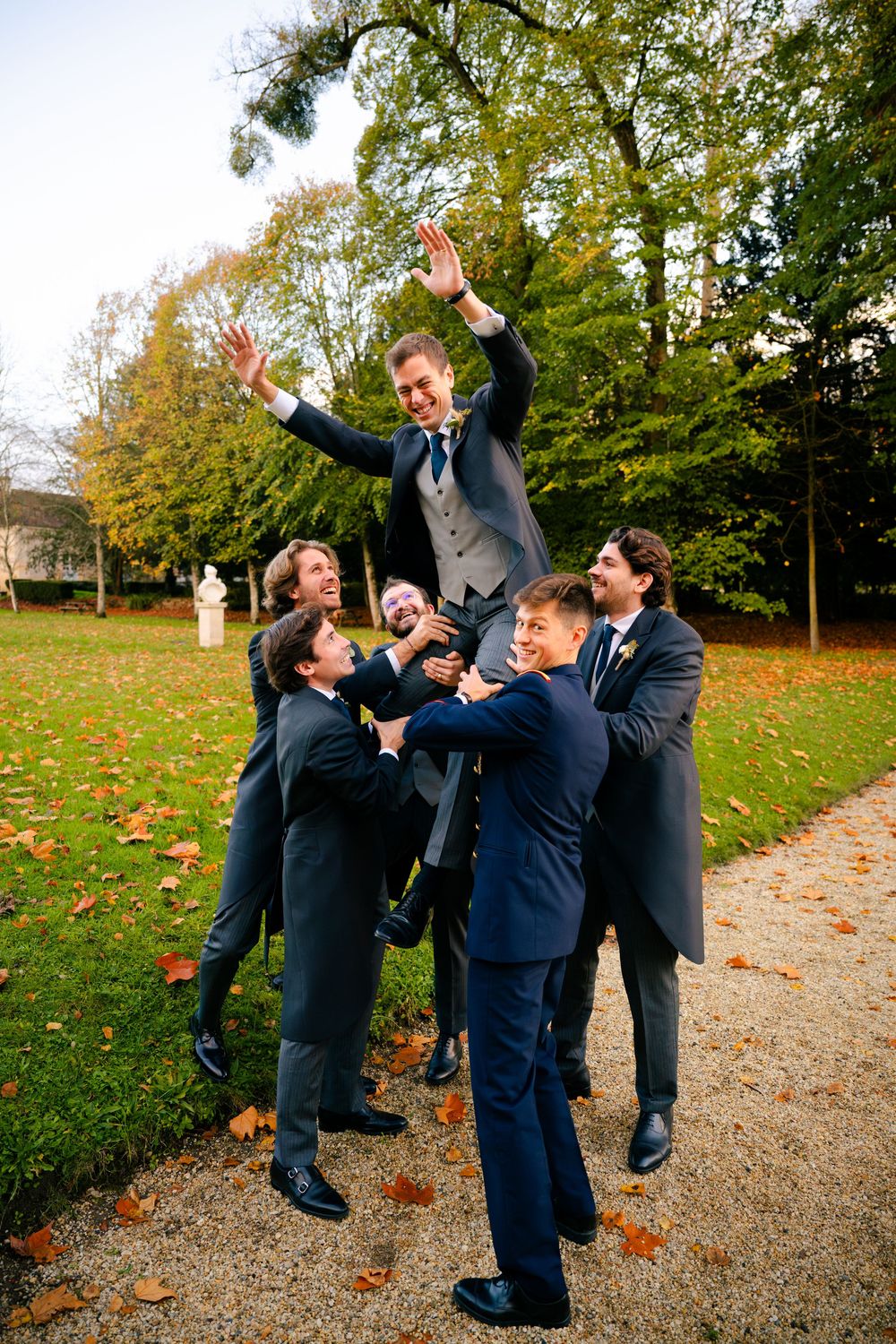 Groomsmen in navy suits playfully lift and celebrate on a fall day with autumn leaves scattered on the ground.