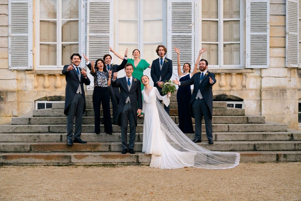 Wedding party stands on stone steps of historic building with shuttered windows and flowing white veil in breeze.